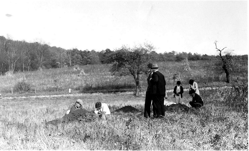 Two men in suits look over the excavation in a field.