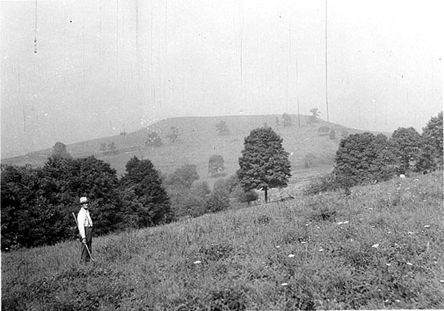 A farmer stands in the foreground and the mesa-like Fort Hill is visible in the distance.
